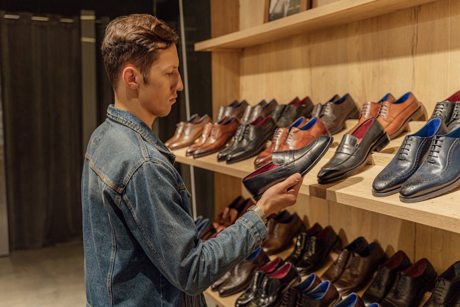 A man in denim jacket selects leather shoes from a store shelf indoors.